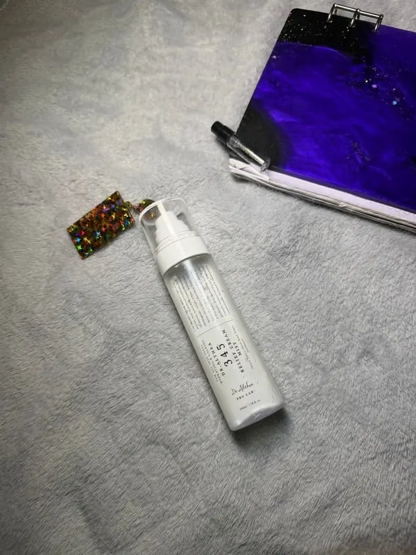 Clear face mist bottle styled on a soft grey throw beside a purple pouch